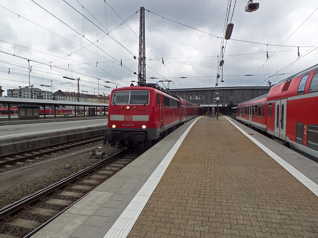 111 227, die 111 mit der höchsten Ordnungsnummer und einem winkenden Lokführer an Bord, verlässt am 29.07.14 München HBF auf der Fahrt nach Pfaffenhofen (Ilm). Auf dem Nebengleis fährt indessen ein weiterer Regionalzug ein, der die aktuell im Süden wohl recht beliebte Nutzung einstöckiger Steuerwagen in Doppelstockzügen demonstriert.