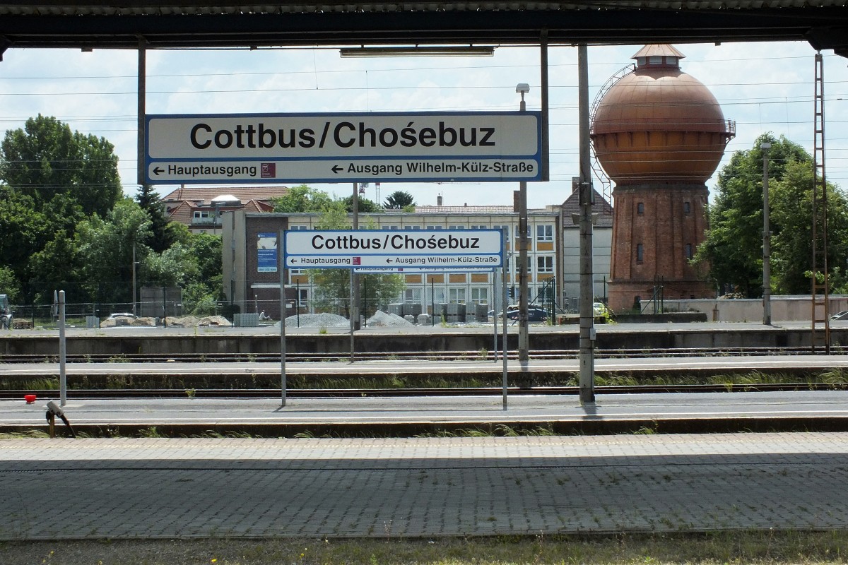 Cottbus. Wir befinden uns im Land der Sorben, neben Friesen, Dänen und Roma eine von vier offiziell anerkannten Nationalen Minderheiten in der Bundesrepublik Deutschland. In weiten Teilen Brandenburgs und Sachsen sind Straßenschilder oder, wie hier zu sehen, Bahnhofsschilder deshalb zweisprachig ausgeführt: Einmal hochdeutsch, einmal sorbisch.
Muttersprachlich gesprochen wird die Sprache heute kaum noch, historisch ist sie aber immer noch bedeutsam genug, um sie allgegenwärtig zu halten. Nicht zuletzt weil Sprache immer eine wichtige Identitätsstiftung war und ist. Bahnhofsszene in Cottbus HBF, wo noch immer die weißen Stationsschilder hängen, die ich sonst in keinem Bahnhof auf meiner Tour sichten konnte, 30.5.14