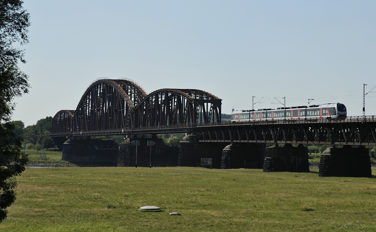 Da die Züge der RB 35 von Gelsenkirchen nach Mönchengladbach wegen Bauarbeiten während der Sommerferien in Nordrhein-Westfalen nicht über Duisburg-Rheinhausen verkehren können, werden sie über die Rheinbrücke in Duisburg-Beeckerwerth umgeleitet. Dadurch ergibt sich die Gelegenheit, sie auf der ansonsten nur vom Güterverkehr genutzten Haus-Knipp-Brücke aufzunehmen. Am 30.07.2020 überquert ein Stadler Flirt 3 von Abellio Rail NRW die Brücke auf der Fahrt nach Gelsenkirchen. Abellio Rail ist ein Tochterunternehmen der niederländischen Staatsbahn. Der Deckel im Vordergrund (Grundwasserbrunnen) gehört zum nahegelegenen Wasserwerk, das aus dem Rheinuferfiltrat Trinkwasser gewinnt.