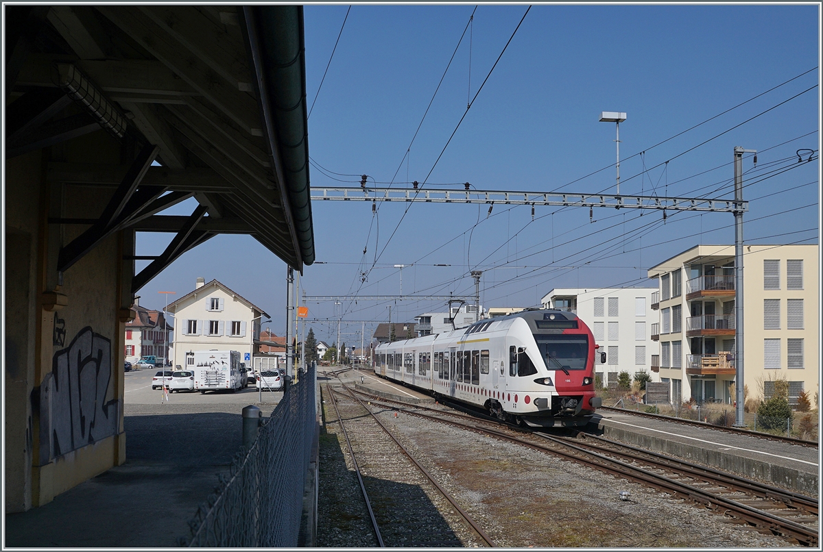 Der Bahnhof von Sugiez zeigt sich bis auf wenige Details wie den Maschengartenzaun an der Güterschuppenrampe und dem P&R zwischen Bahnhof und Güterschuppen noch weitgehend im Zustand wie zahlreiche Landbahnhöfe sich vor gut dreissig, vierzig Jahren präsentierten. Doch Umbauten sind schon geplant....
Der TPF RABe 527 195 verlässt Sugiez in Richtung Romont.

9. März 2022