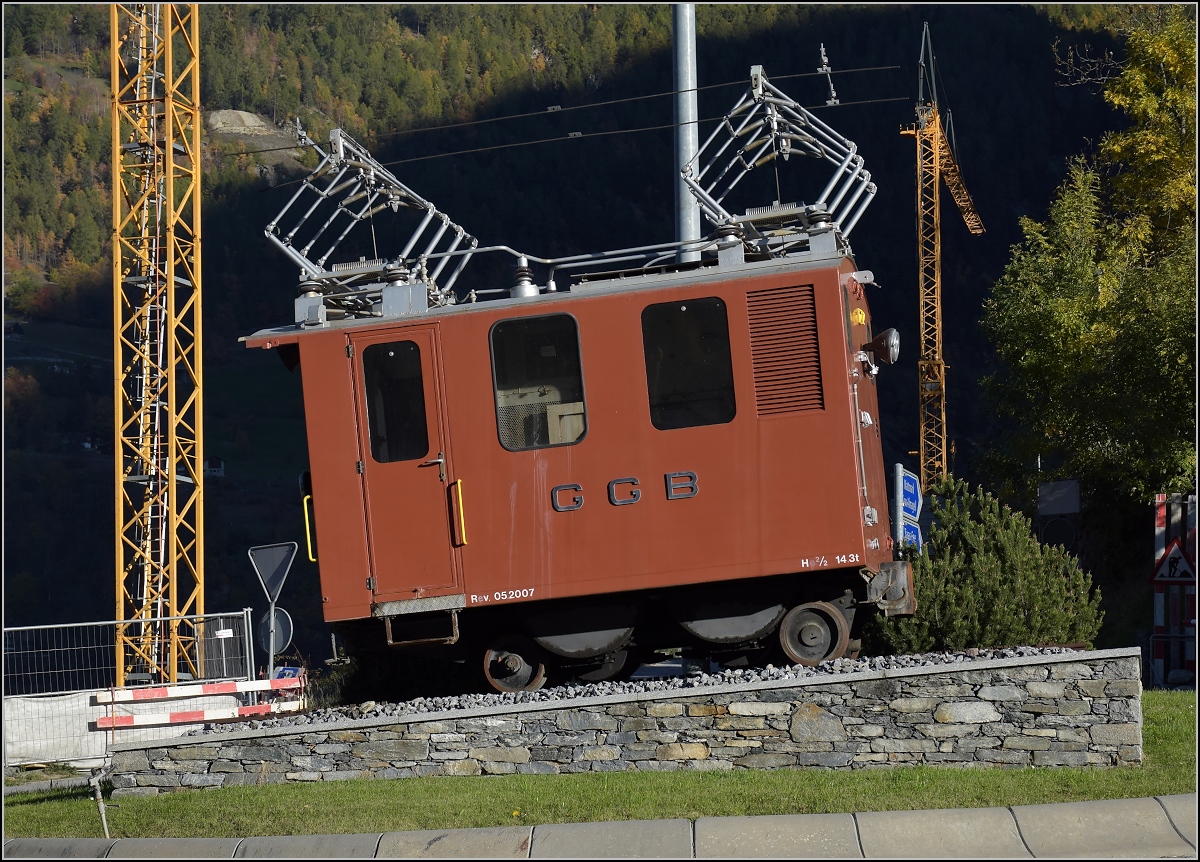 Die Lokomotive He 2/2 14 der Gornergratbahn erwartet den Autofahrer schon an der Weggabelung zwischen Saastal und Mattertal. Mittem im Kreisverkehr kann man schon sehen, was einen früher erwartet hätte. Ein Stück eigenen Fahrdraht hat die kleine Drehstromlok schon mitgebracht, interessant die zweipoligen Stromabnehmer. Stalden, Oktober 2017.