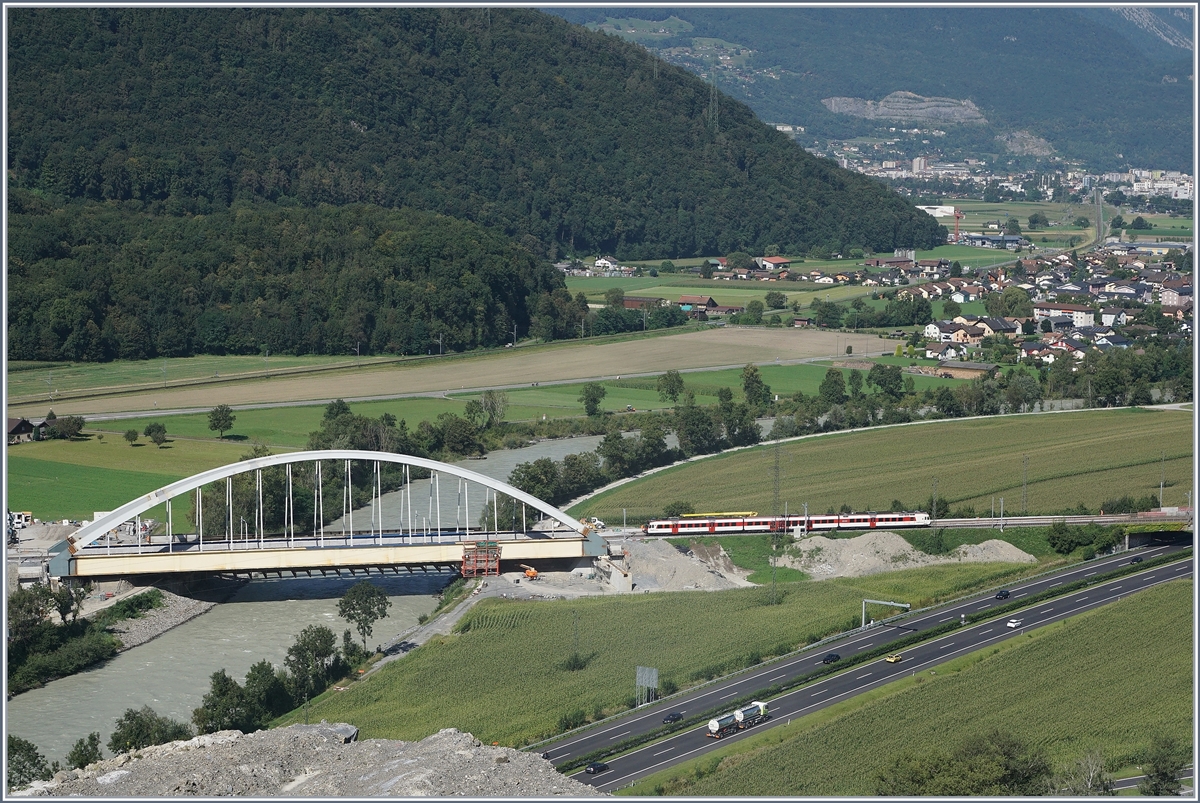 Die Suche nach einem besseren Fotostelle für die neue Rhonebrücke führten mich an Nahe an den sicheren Abgrund. Abschrankungen des Kieswerkes beachtend und so auf öffentlichem Grund bleibend, hat man einen ungefährlichen und schönen Blick auf die neuen Rhonebrücke, die demnächst an ihren definitiven Standort verschoben werden wird. Auf dem Bild erreicht ein  Walliser Domino  die Brücke, welcher für die seit Fahrplanwechsel teilweise fehlenden Halte der IR als RE zwischen St-Maurice und Aigle verkehrt. 26. August 2016 