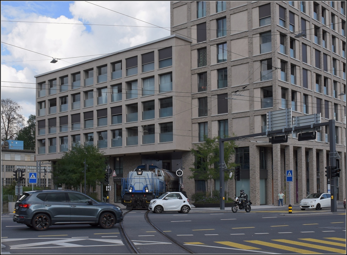 Gravita Am 847 003 'Littau' schiebt einen schweren Stahlzug von der Emmenwaid durch das Eckhaus in den Bahnhof Emmenbrücke. Die Einfahrt nach Emmenbrücke ist noch nicht frei. Und irgendwie haben selbst Kleinstwagen offenbar schon SUV-Dimensionen, wie der Vergleich des Zweisitzers mit der Diesellok erahnen lässt. August 2021. 