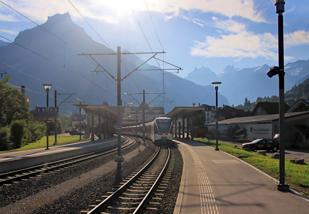 Zentralbahn, Strecke Luzern - Engelberg: Im grellen morgendlichen Gegenlicht am Fusse einer imposanten Berglandschaft steht ein Zug in Engelberg bereit für die Rückfahrt nach Luzern. Dreiteiliger Steuerwagen ABt 941 zuvorderst. 12.August 2024 