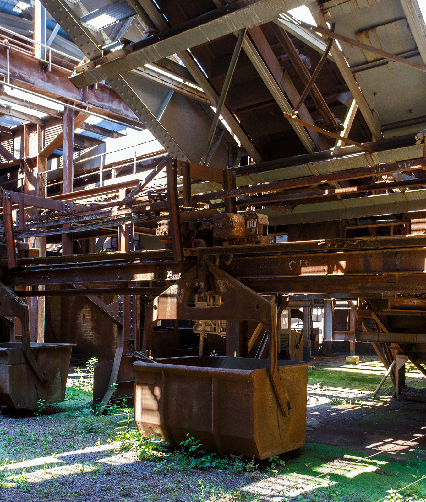 Auch eine Eisenbahn (Schwebebahn): Die H�ngebahn der V�lklinger H�tte am 01 Juli 2025, hier einer der selbstfahrenden H�ngebahnwagen unter der M�llerhalle. 

Die H�ngebahn ist eine Einschienenbahn bzw. Schwebebahn mit selbstfahrende H�ngebahnwagen, bei den Schr�gaufz�gen auf die Gichtb�hne wurden sie mit Seilz�gen unterst�tzt. 

Die H�ngebahn war seit 1911 in Betrieb. 1985 fuhren pro Stunde 240 beladene H�ngebahnwagen zur  Gicht . Sie liefen �ber die sogenannte Gichtb�hne in einer H�he von 27 Metern, die Rohstoffe (Eisenerz, Sinter, Schrott, Kalk und Koks) um die 6 Hoch�fen von dort zu beschicken. Die Gleisl�nge der H�ngebahn betrug �ber 6 km.

Der M�ller (Eisenerz, Sinter, Schrott und Kalk wurden in der M�llerhalle (s�d�stlich der Hoch�fen) in selbstfahrende H�ngebahnwagen verladen und �ber einen vierspurigen Schr�gaufzug auf die Gichtb�hne gezogen. Auf der Gichtb�hne laufen mehrere H�ngebahnspuren an den sechs Hoch�fen vorbei und wieder zur�ck zum Schr�gaufzug, mit kreisf�rmigen Abzweigschleifen um jeden Hochofen herum. Auf der Nordwestseite der Gichtb�hne l�uft ein kleinerer zweispuriger Schr�gaufzug hinunter zur Kokerei, um den Koks f�r die Hoch�fen zu f�rdern.

Die Gichtb�hne verbindet alle sechs Hoch�fen mit einer Ebene. Auf dieser Ebene werden die H�ngebahnwagen entlanggef�hrt. Die Gichtb�hne ist ca. 240 Meter lang und erstreckt sich in einer H�he von exakt 27 Meter.  Die Gichtb�hne hat Ihren Namen von dem Wort  Begichten , was das Bef�llen der Hoch�fen mit den Rohstoffen f�r die Eisenerzeugung bezeichnet. Eisenerz, Sinter, Zusatzstoffe, Koks, aber auch eisenhaltige Reststoffe aus der Industrie oder Schrott wurden in die Gicht, den Einf�lltrichter des Hochofens, gekippt. Die „Haube/obere Glocke“, der obere Verschluss des Hochofens, wurde angehoben, dann f�nf H�ngebahnwagen um den Trichter gefahren. Unterhalb der Haube befindet sich ein zweiter Verschluss, die „Gicht-Glocke“. Sie blieb geschlossen, um das Gichtgas aufzufangen. Das Material wurde auf die Glocke gekippt, dann die Haube abgesenkt. Erst wenn die Haube geschlossen war, wurde die Glocke nach unten gefahren: Das Material rutschte in den Hochofenschacht. Diese Verschlussart nannte man „Doppelglockenverschluss“, bei modernen Hoch�fen werden heute glockenlose Gichtverschl�sse verwendet. 
