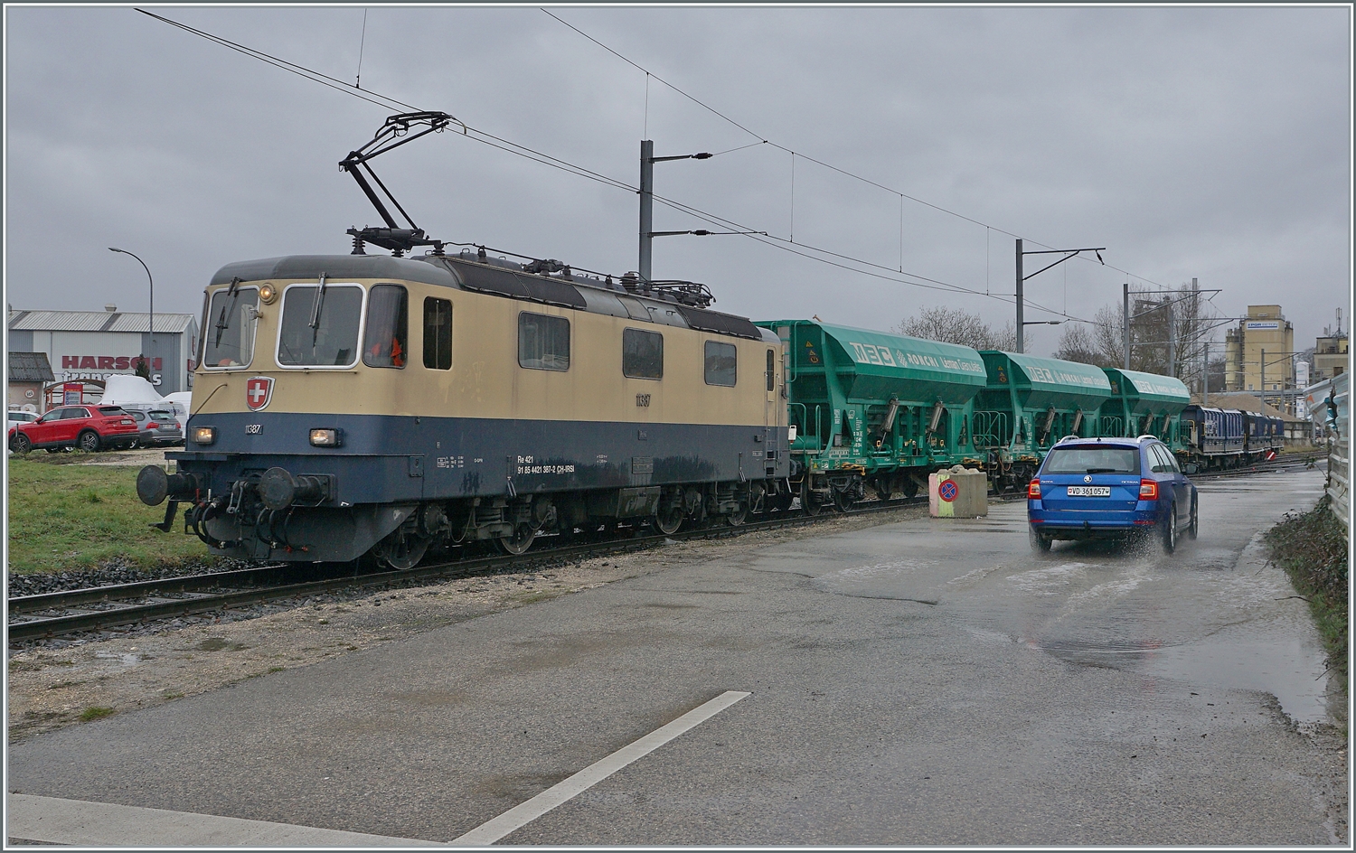 Bei sehr schlechtem Wetter ist die Die IRSI/IGE  Rheingold  Re 4/4 II 11387 (Re 421 387-2) kommt mit ihrem Güterzug auf dem Industriegleis in Gland auf der Fahrt vom Kieswerk zum Bahnhof. 
Noch kurz zum  schlechten Wetter : der Vorteil, dass kein Gegenlicht herrschte wurde mit dem Nachteilen der mässigen Lichtes und der Wassertropfen auf dem Objektiv erkauft. 

22. Februar 2024