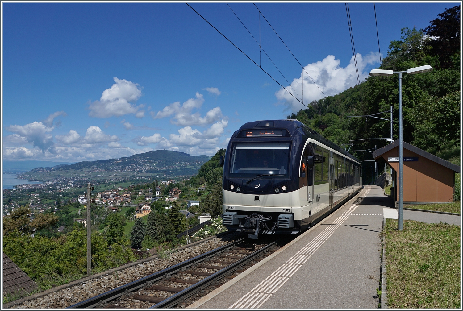 Bild mit Aussicht: Während der CEV MVR ABeh 2/6 7503   Blonay-Chamby  als Regionalzug von Montreux nach Les Avants den Bahnhof  Sonzier erreicht, gleitet links der Blickj weit über die Riviera Vaudoise und das Lavaux. 

28. Mai 2024