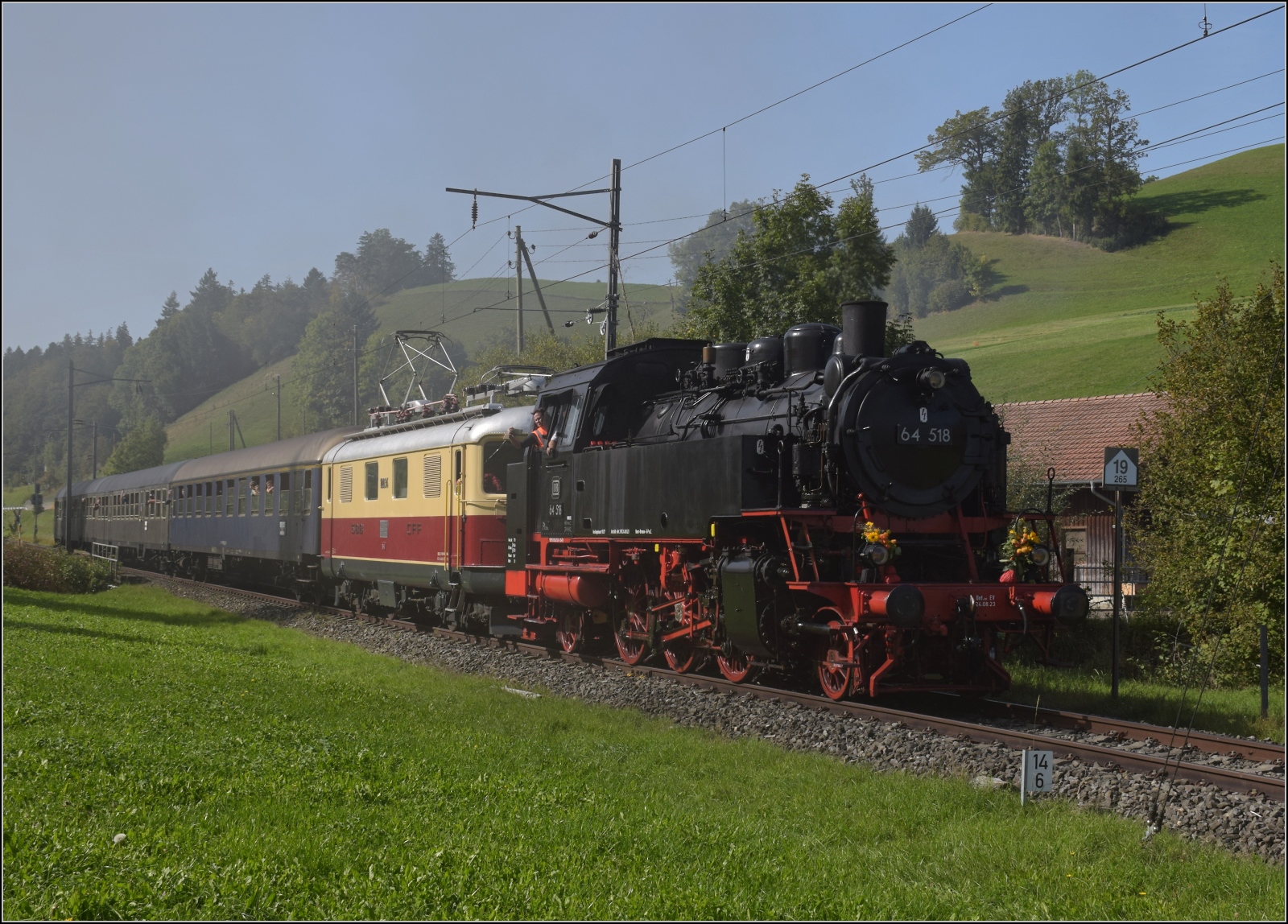 Dampftage Huttwil mit frisch genesenem Bubikopf. 

64 518 und Re 4/4 I 10034 mit zwei Silberlingen und einem erstklassigen Schnellzugwagen der DB kommt in Dürrenroth an. Rauch und Dunst stammen aber nicht von ihr selbst, sondern die im Bahnhof wartende Ec 3/4 2 der Solothurn-Münster-Bahn sorgte als Nebelmaschine für Stimmung. Oktober 2023