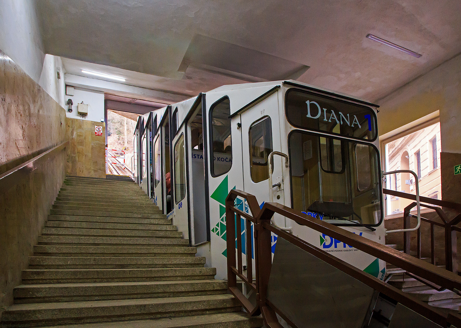 Der Wagen „DIANA 1“ der Standseilbahn Diana (tschechisch Lanová dráha Diana) in Karlsbad (Karlovy Vary) am 18.04.2023 in der Talstation Stará louka (Alte Wiese).

Die Stadtseilbahnen (Diana sowie Imperial) stellen in Karlsbad schon seit Anfang des vergangenen Jahrhunderts ein traditionelles und beliebtes Verkehrsmittel dar. Sie gehören inzwischen zum Stadtbild und sind ein beliebtes Touristenziel.

Diana-Standseilbahn:
Die Standseilbahn Diana (tschechisch Lanová dráha Diana) wurde im August 1912 eröffnet und beginnt an der Station Alte Wiese (Stará louka) auf einer Höhe von 389 m und führt auf die Freundschaftshöhe 556 m über NN, wo sich der Diana-Aussichtsturm befindet. Die Talstation befindet sich gleich neben dem Grandhotel Pupp in der Gasse Mariánská. Den Höhenunterschied von 167 m überwindet sie auf einer Strecke von 437 m. Im Bereich der abtsche Ausweiche liegt auch die Mittelstation Jelení skok (Hirschsprung) in 473 m Höhe. Die maximale Steigung beträgt 432,3 ‰, durchschnittlich liegt diese bei 398,1 ‰. Die Bahn wurde insgesamt dreimal renoviert, zwischen 1963 und 1965, 1972 und noch einmal zwischen 1984 und 1988. Im Gegensatz zur Bahn zum Personennahverkehrs der Stadt. Auch sie wird im 15-Minuten-Takt befahren und benötigt 4 Minuten für eine Fahrt bei einer Geschwindigkeit von 2,04 m/s. Die Gleise sind vom Typ A, die Spurweite beträgt 1.000 mm. Die leer 7.900 kg wiegenden Wagen sind für 49 + 1 Fahrer ausgelegt. Das Seil hat einen Durchmesser von 28 mm. Die Antriebsleistung beträgt 51 kW, die Antriebsregulation erfolgt mit Hilfe von Thyristoren, 

Die Seilbahn ist ein Projekt des Ing. H. H. Peter aus Zürich. Der Bau der Seilbahn wurde von der Wiener Baufirma Leo Arnoldi mit Hilfe von lokalen Handwerkern und Firmen durchgeführt. Die erste Generalüberholung der Seilbahn geschah in den Jahren 1963-1965, als die Seilbahnen neue Metallwagons aus der Produktion des Tatra Smíchov Wagon bekamen. Die Rekonstruktion der elektrischen Ausrüstung wurde 1972-73 durchgeführt.Es ist ein traditionelles und sehr beliebtes Verkehrsmittel auf der Freundschaftshöhe in Karlsbad, das hauptsächlich von den Touristen der Stadt genutzt wird. Von der Talstation wird man zum Gipfel gebracht, wo sich der Aussichtsturm Diana und das Wald-Restaurant und Café Diana befindet.

Betreiber der Seilbahnen ist die DPKV - Dopravní podnik Karlovy Vary a. s. (Verkehrsbetrieb Karlsbad AG). Der Verkehrsbetrieb Karlsbad AG ist ein traditioneller Betreiber des öffentlichen Verkehrs in Karlsbad. Er betreibt tagsüber und auch in den Nachtstunden die regelmäßigen Stadtbusverkehrslinien, Sonderverkehrslinien und Vorortverkehrslinien. Ein Besonderheit des Karlsbader ÖÖV ist der Verkehr der Stadtseilbahn vom Theaterplatz (Divadelní náměstí) zum Hotel Imperial und vom Grandhotel Pupp zum Aussichtsturm Diana. Der Verkehrsbetrieb Karlsbad AG bietet der Öffentlichkeit auch weitere, überwiegend mit der Beförderung zusammenhängende Dienstleistungen an. 

DATEN der Seilbahn DIANA:
Höhenunterschied: 167 m
Streckenlänge:  453 m
Spurweite: 1000 mm (Meterspur)
Maximale Steigung:  432,3 ‰
Leergewicht eines Wagens: 7.900 kg
Kapazität eines Wagens: 49 + 1 Fahrer
Seildurchmesser: 28 mm
Antriebsleistung: 51 kW
Höchstgeschwindigkeit: 7,3 km/h
Taktung: alle 15 Minuten
Fahrtdauer: ca. 3 Minuten
Preis (z.Z.) für Hin- und Rückfahrt: 150 CZK