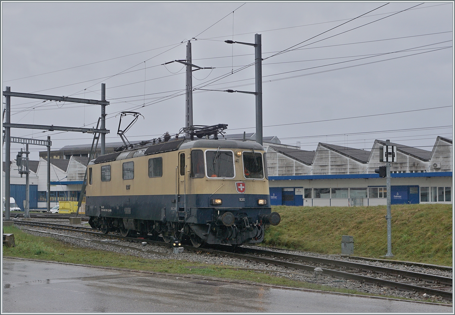 Die von Morges gekommene IRSI Re 4/4 11387 verlässt in Gland die Strecke nach Genève und fährt in über ein Industriegleis in Richtung Kieswerk um dort ihren Zug nach Apples abzuholen. Die angemietete IRSI/IGE Re 4/4 II 11387 (Re 421 387-2) ersetzt die zur Zeit in der Revision in Bellinzona weilende BAM MBC MBC Re 420 506.

22. Feb. 2024