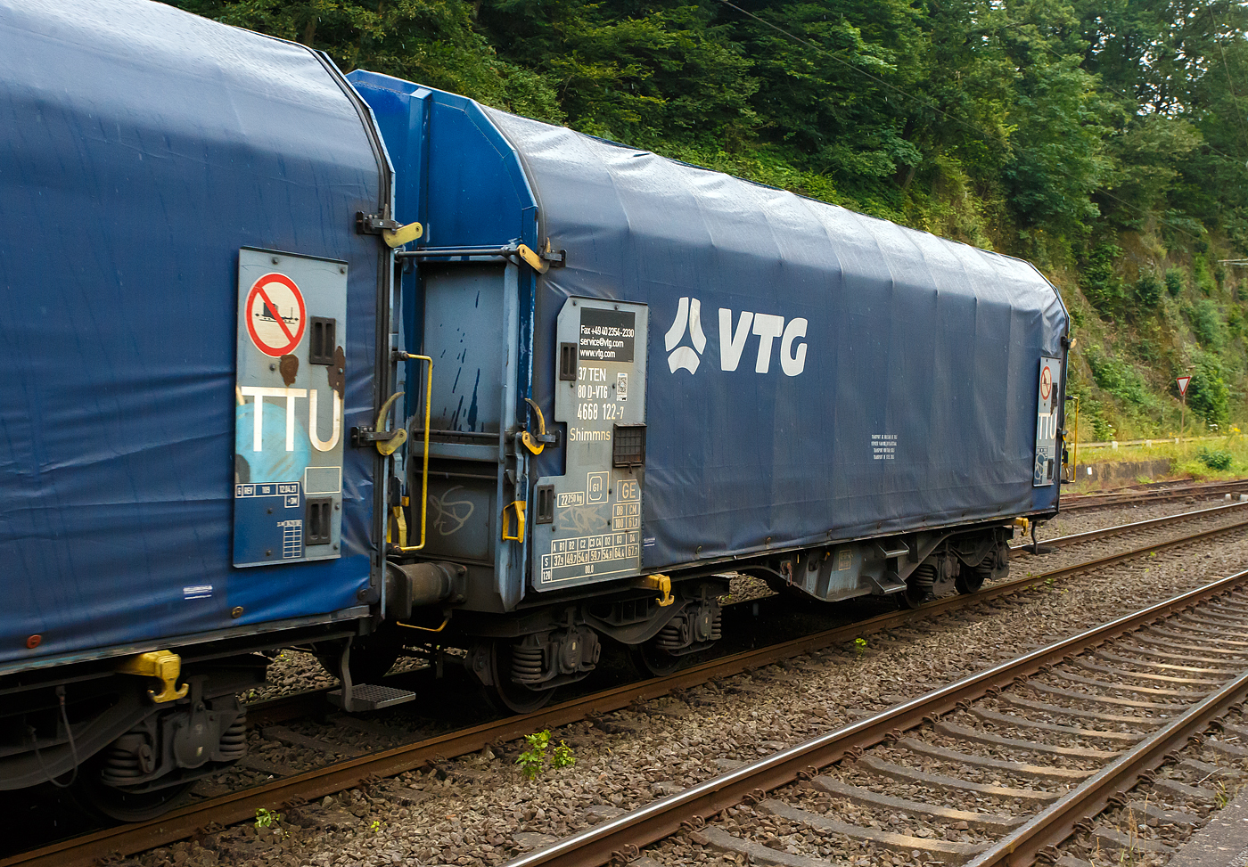 Vierachsiger Drehgestell-Flachwagen mit verschiebbarem Planenverdeck und Lademulden für Coiltransporte, 37 80 4668 122-7 D-VTG, der Gattung Shimmns-ttu (VTG Bez. F46.520D), der VTG Rail Europe GmbH (Hamburg), am 03 Juli 2024 eigereiht im Zugverband (Zudschluss) der KSW bei einer in Zugdurchfahrt im Bf Scheuerfeld/Sieg, in Richtung Herdorf via Betzdorf.

Diese Wagen sind speziell für den Transport witterungsempfindlicher Blechrollen (Coils) bestimmt. Er besitzt feste Stirnwände, 5 fest im Untergestell eingebaute Lademulden. Die Wagenabdeckung erfolgt mit einer PVC-beschichteten Gewebeplane, die auf 11 Rohrrahmen aufliegt. Über Laufwagen ist das Planendach in Wagenlängsrichtung verschiebbar. Die Verriegelung des Planendaches an den Stirnwänden erfolgt über eine 4-Punkt-Zentralverriegelung, die sowohl vom Erdboden als auch von der Rampe aus bedient werden kann. Das Planenverdeck lässt sich an einem Wagenende so weit zusammenschieben, dass ca. 2/3 der Ladefläche zur Beladung freigegeben wird. Hierdurch ist eine einfache Beladung des Wagens sowohl von oben als auch von der Seite aus möglich.

TECHNISCHE DATEN:
Spurweite: 1.435 mm
Achsanzahl: 4 (in 2 Drehgestelle)
Länge über Puffer: 12.040 mm
Länge der Ladefläche: 10.800 mm
Drehzapfenabstand: 7.000 mm
Achsabstand im Drehgestell: 1.800 mm
Eigengewicht: 22.250 kg
Höchstgeschwindigkeit: 100 km/h (beladen) / 120 km/h (leer)
Maximales Ladegewicht: 67,7 t (ab Streckenklasse D4), DB CM 61,7 t
Mulden: 5 Stück, im Untergestell integriert
Werkstoff Muldenmatten: Tecthan
Festlegearme: 20 Stück, stufenlos verstellbar, ausgestattet mit Schutzplatten
Coil-Breite: min. 342 mm bis max.  2.042 mm
Kleinster bef. Gleisbogenradius: 35 m
Bauart der Bremse: KE-GP-A (K)
Bremssohle: C 810
Intern. Verwendungsfähigkeit: TEN-GE